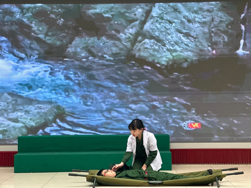 A person in green uniform lying on a green blanket with a large screen behind herDescription automatically generated
